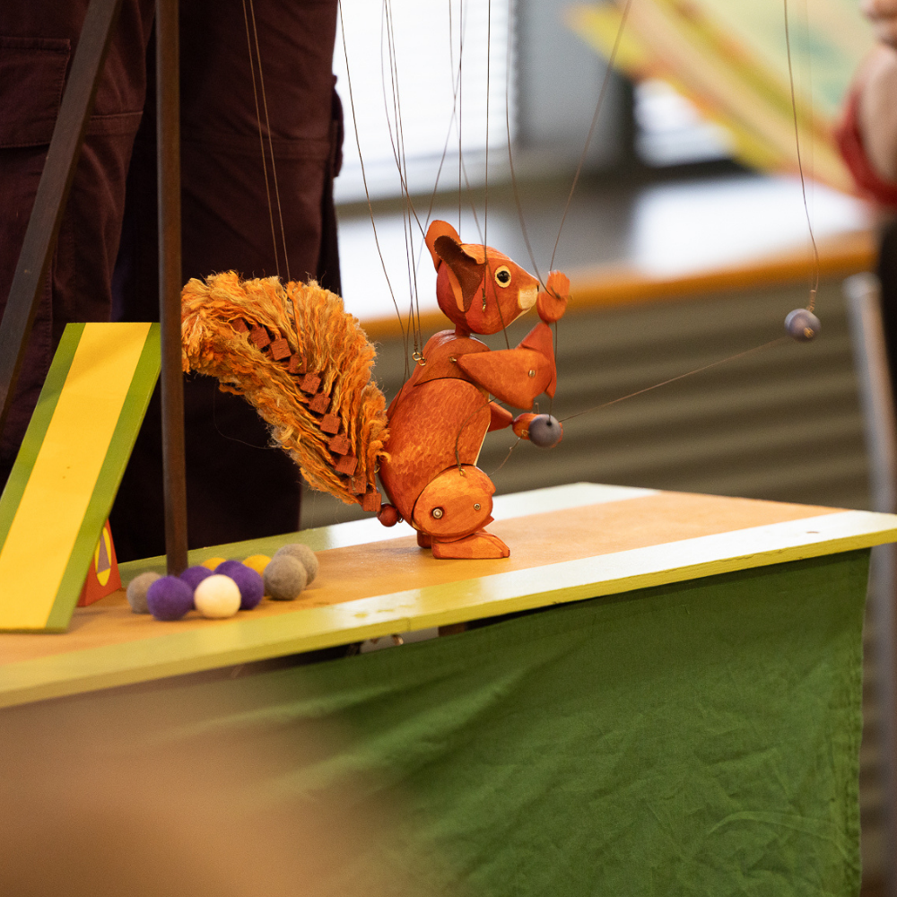 A wooden squirrel marionette being performed on a small stage during a Libraries Unlimited puppet show, with colourful props and craft materials around the setup.