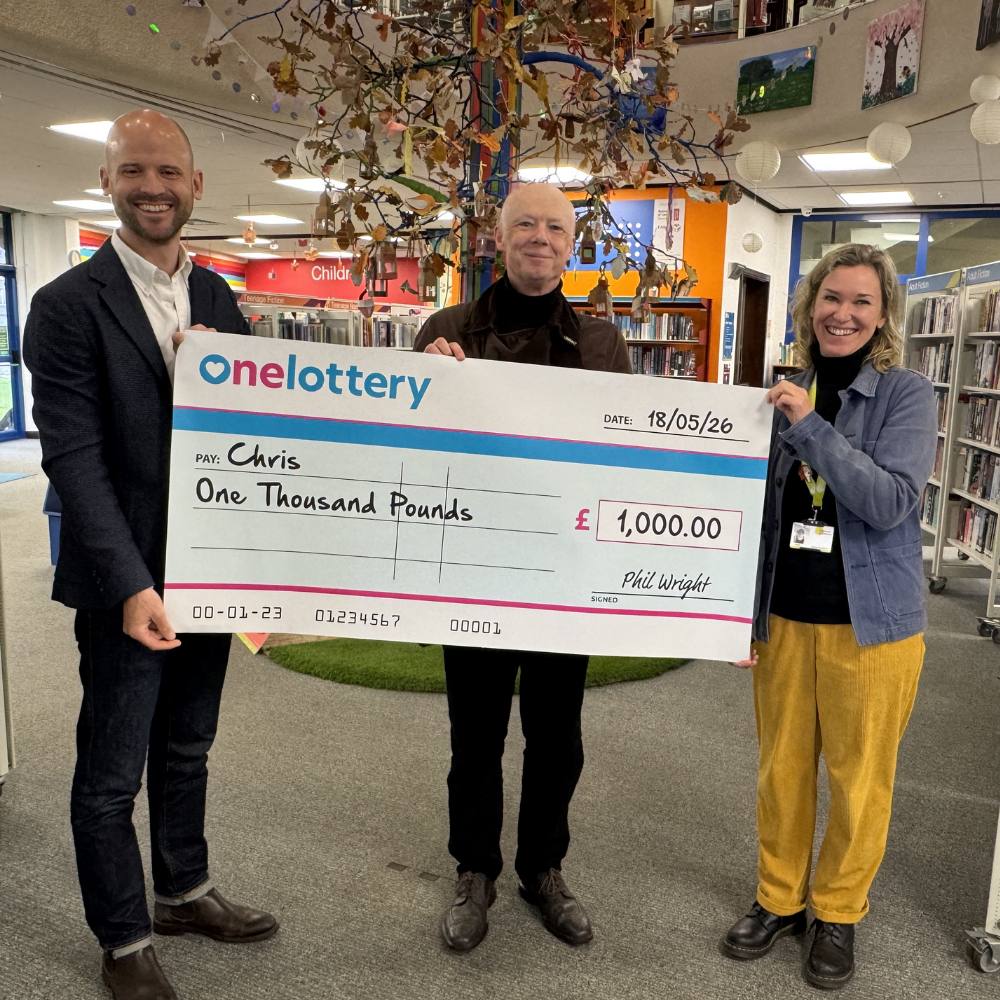 Three people stand in a library holding a giant check made out to Chris for £1,000