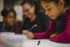 Mother and two daughters attending arts workshop, shown drawing and writing