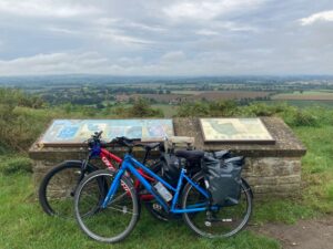 Two bikes, one red and one blue, lean on a low stone wall. Overlooking fields